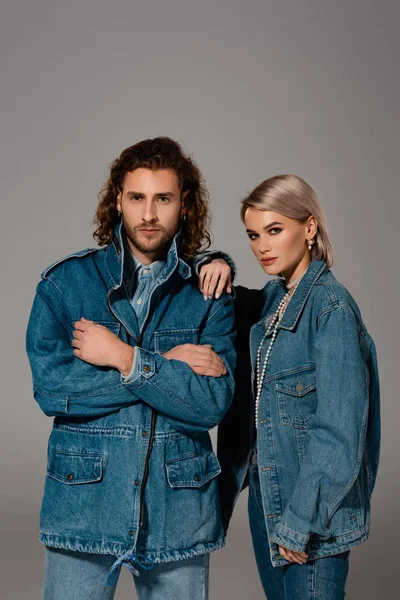 Homem elegante com braços cruzados e mulher em jaqueta jeans olhando para a câmera isolada em cinza — Fotografia de Stock