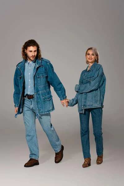 Стильный мужчина и улыбающаяся женщина в джинсовых куртках держась за руки на сером фоне — стоковое фото