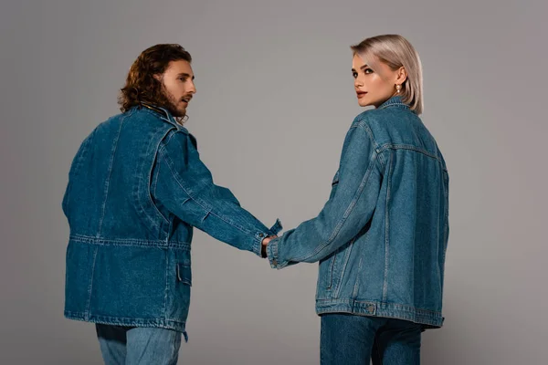 Vista posteriore di uomo e donna alla moda in giubbotti di jeans che tengono le mani isolate sul grigio — Foto stock