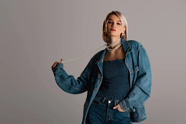 Attraktive Frau in Jeansjacke mit Perlenkette und Blick in die Kamera vereinzelt auf grau — Stockfoto