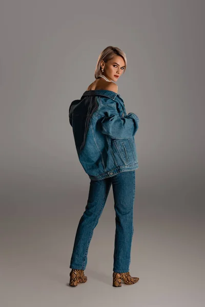 Attraktive Frau in Jeansjacke und Jeans blickt in die Kamera auf grauem Hintergrund — Stockfoto