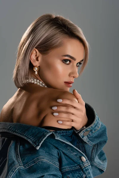 Atractiva mujer en chaqueta de mezclilla con collar de perlas mirando hacia otro lado aislado en gris — Stock Photo