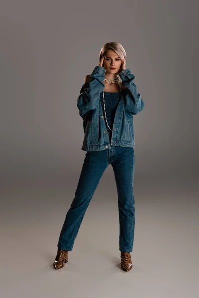 Привлекательная и уставшая женщина в джинсовой куртке и джинсах трогательная голова на сером фоне — стоковое фото