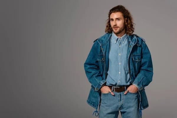 Hombre guapo y elegante en chaqueta de mezclilla y jeans con manos en bolsillos aislados en gris - foto de stock