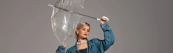 Plano panorámico de mujer atractiva en chaqueta de mezclilla sosteniendo paraguas aislado en gris - foto de stock