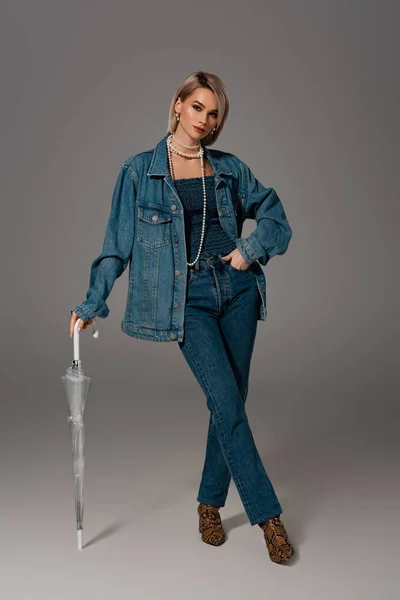 Attractive woman in denim jacket and jeans holding umbrella on grey background — Stock Photo