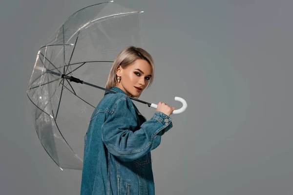 Attractive woman in denim jacket holding umbrella isolated on grey — Stock Photo