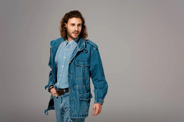 Красивый и стильный мужчина в джинсовой куртке с рукой в кармане, изолированный на сером — стоковое фото