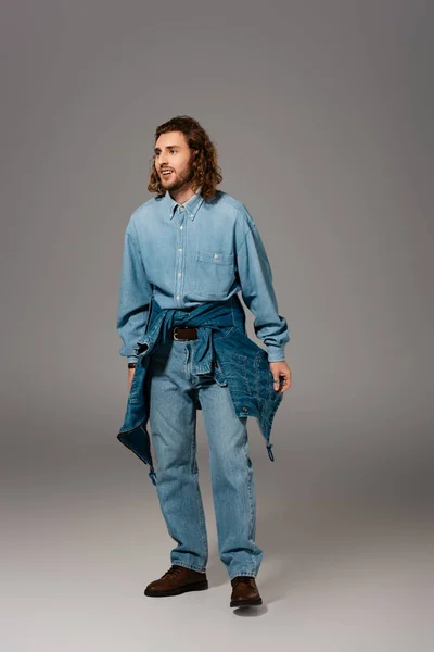 Hombre sonriente en camisa de mezclilla y jeans mirando hacia otro lado sobre fondo gris — Stock Photo