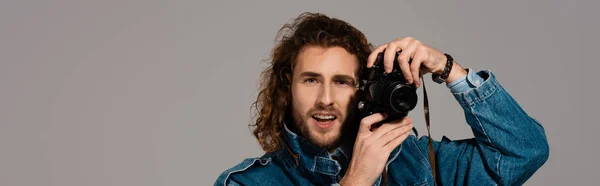 Panoramic shot of smiling man in denim jacket holding digital camera isolated on grey — Stock Photo