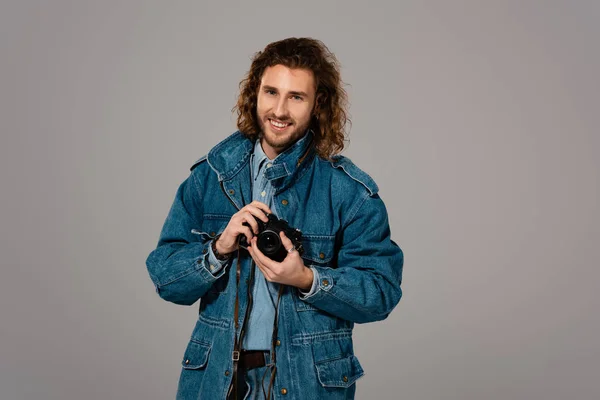 Homem bonito e sorridente em jaqueta de ganga e jeans segurando câmera digital isolada em cinza — Fotografia de Stock