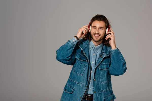 Uomo sorridente in giacca di denim che ascolta musica con cuffie isolate su grigio — Foto stock
