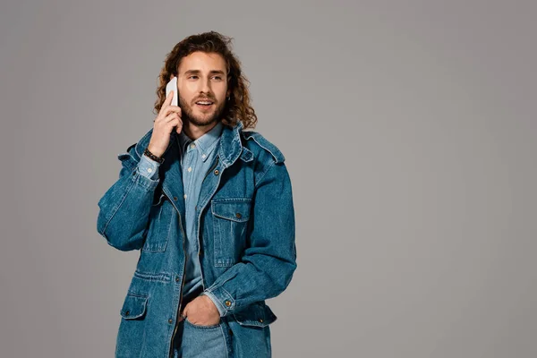 Hombre guapo y elegante en chaqueta de mezclilla hablando en el teléfono inteligente aislado en gris - foto de stock