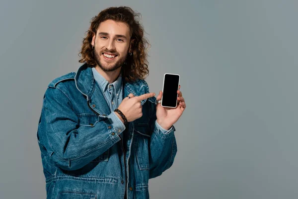 Homem sorridente em jaqueta de ganga apontando com o dedo para smartphone isolado em cinza — Fotografia de Stock