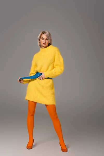 Attraktive Frau im gelben Kleid berührt Gürtel und blickt in die Kamera auf grauem Hintergrund — Stockfoto