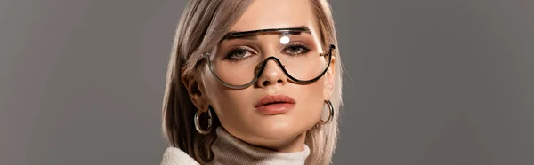 Панорамний знімок привабливої жінки в окулярах і сережках дивиться на камеру ізольовано на сірому — стокове фото