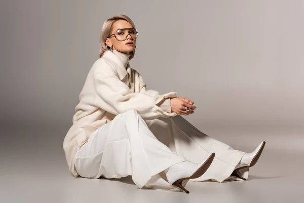 Atractiva mujer de abrigo blanco y pantalones sentados sobre fondo gris - foto de stock