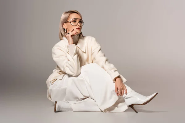 Mujer atractiva y pensativa en abrigo blanco y pantalones sentados sobre fondo gris - foto de stock