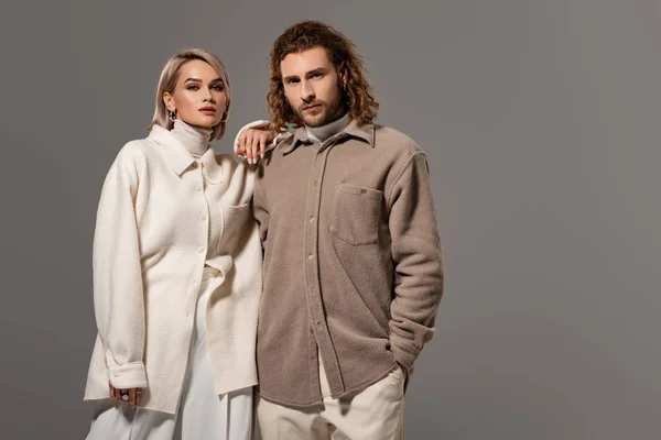 Frau in weißem Mantel und Mann im Hemd mit der Hand in der Tasche schauen vereinzelt in die Kamera auf grau — Stockfoto