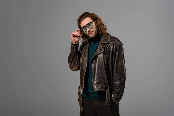 Handsome man in leather jacket touching glasses and looking at camera isolated on grey — Stock Photo
