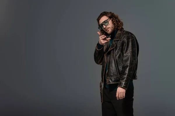 Handsome man in leather jacket looking at camera and showing gun gesture isolated on grey — Stock Photo