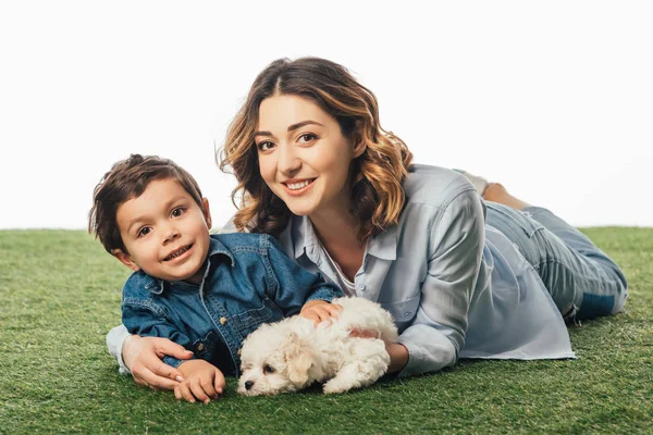 Madre e figlio sorridenti sdraiati sull'erba con cucciolo avanese isolato su bianco — Foto stock