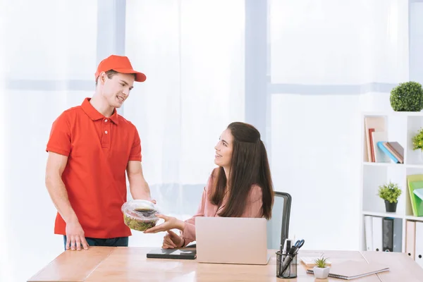 Delivery man giving takeaway salad to smiling businesswoman in office — Stock Photo