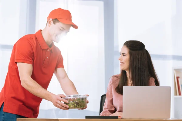 Lächelnder Kurier übergibt Container mit Salat an lächelnde Geschäftsfrau am Tisch — Stockfoto