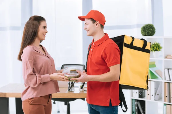 Vista lateral del repartidor hombre con mochila termo dando ensalada para llevar a la mujer de negocios sonriente en la oficina - foto de stock