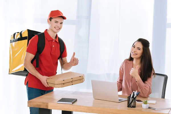 Entrega homem com caixas de pizza mostrando polegares para cima gesto perto de empresária na mesa — Fotografia de Stock