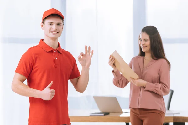 Selective focus of courier smiling at camera and showing like and okay gestures near businesswoman with envelope in office — Stock Photo