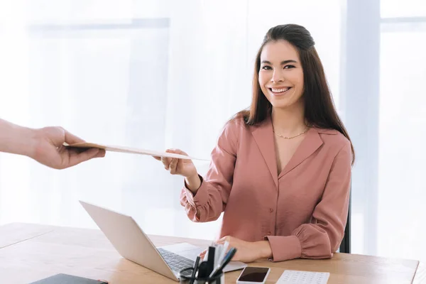 Empresária atraente sorrindo para a câmera perto do correio com envelope no escritório — Fotografia de Stock