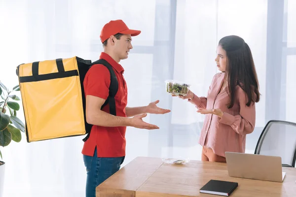 Side view of confused businesswoman with salad in container looking at courier with thermo backpack in office — Stock Photo