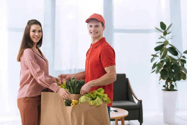 Vista lateral del mensajero sonriendo a la cámara y dando paquetes con verduras frescas a mujer atractiva - foto de stock