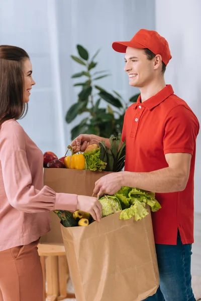 Вид збоку усміхненого кур'єра, що дає пакети зі свіжими фруктами та овочами жінці вдома — стокове фото