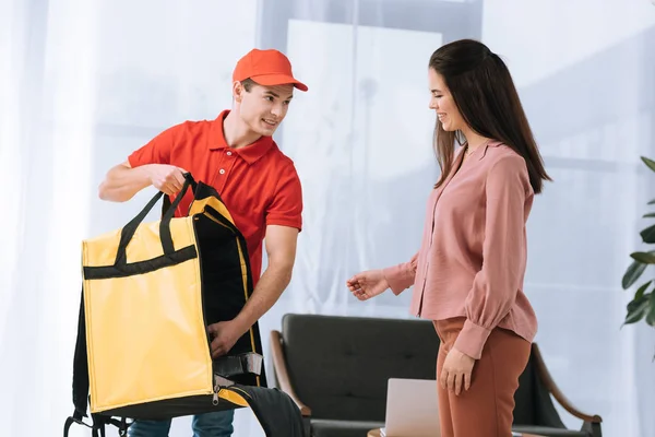 Mensajero sonriente con bolsa de termo cerca de mujer atractiva en casa - foto de stock