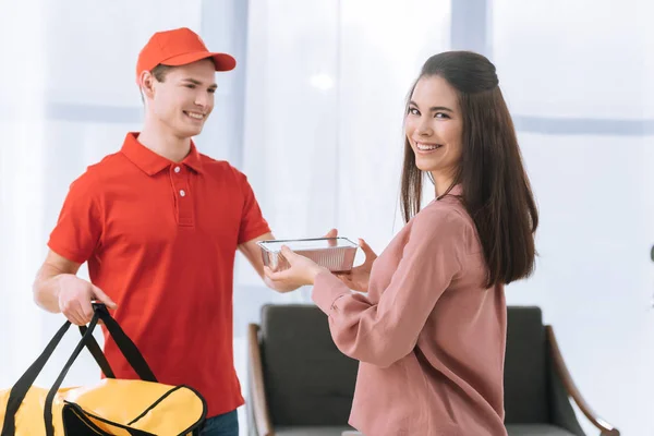 Hermosa mujer sonriendo a la cámara cerca de mensajero con contenedor de alimentos - foto de stock