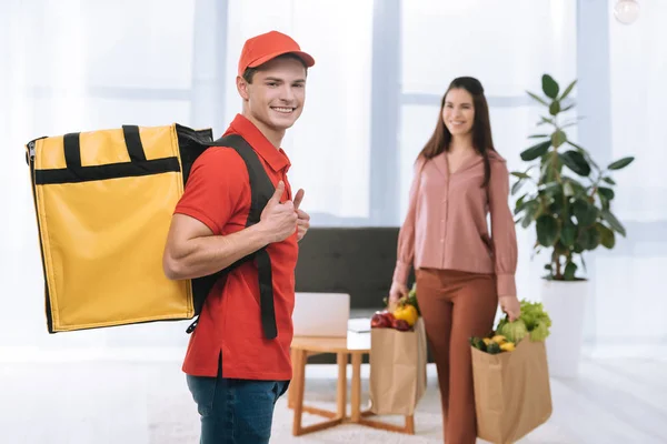 Concentration sélective du courrier souriant avec sac à dos thermique montrant pouces vers le haut près de la femme avec des paquets de légumes frais à la maison — Photo de stock