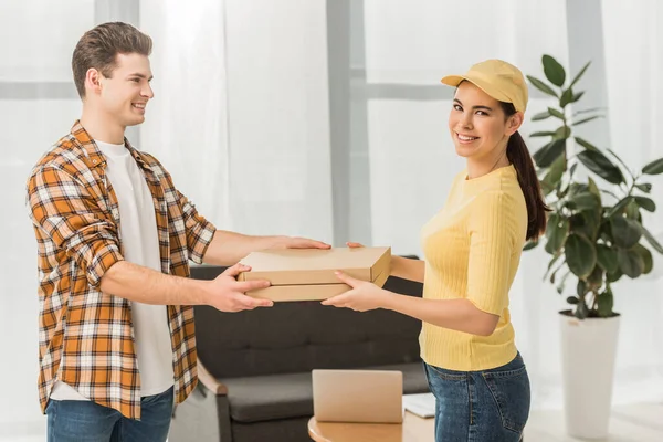 Vista lateral do mensageiro atraente sorrindo para a câmera enquanto dá caixas de pizza para o homem — Fotografia de Stock