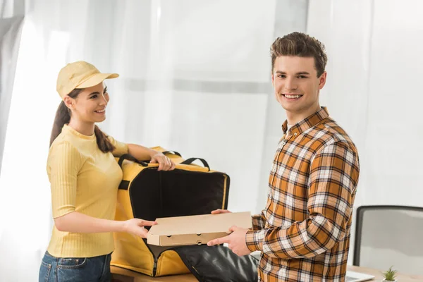 Empresário sorridente com caixa de pizza sorrindo para a câmera perto do correio com saco térmico — Fotografia de Stock