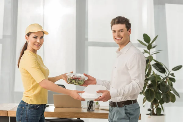 Side view of courier giving food containers to businessman and smiling at camera in office — Stock Photo