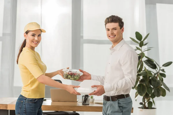 Seitenansicht des Kuriers, der Geschäftsleuten Lebensmittelbehälter schenkt und im Büro in die Kamera lächelt — Stockfoto