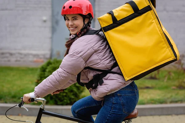 Seitenansicht eines lächelnden Kuriers mit Thermo-Rucksack, der auf der Straße mit dem Fahrrad in die Kamera schaut — Stockfoto