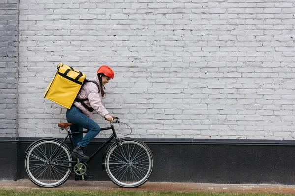 Vista lateral del mensajero sonriente con la mochila termo montar en bicicleta cerca del edificio - foto de stock