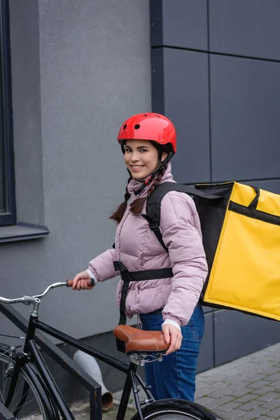 Atractivo mensajero con mochila térmica sonriendo a la cámara cerca de la bicicleta en la calle urbana - foto de stock