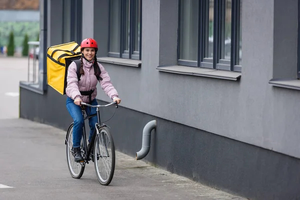 Mensajero sonriente con mochila termo montar en bicicleta cerca del edificio - foto de stock