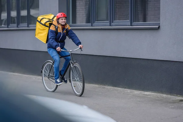 Enfoque selectivo del repartidor sonriente con la mochila termo montar en bicicleta cerca del edificio - foto de stock