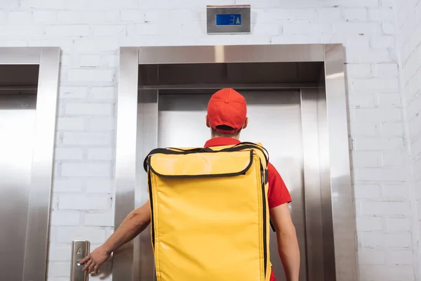 Vista posteriore del corriere in uniforme rossa con zaino termo premendo il pulsante dell'ascensore — Foto stock