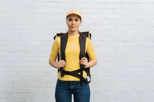 Attractive courier in yellow uniform with thermal backpack looking at camera near brick wall — Stock Photo