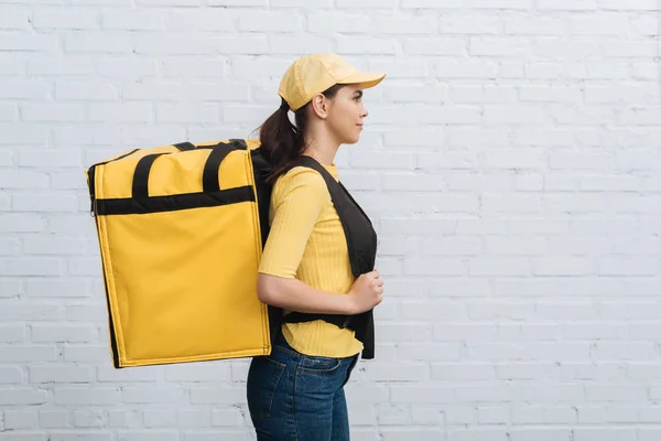 Vista lateral del mensajero en uniforme amarillo con mochila térmica de pie cerca de la pared de ladrillo - foto de stock